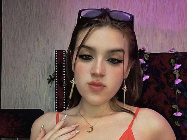 ZoeyClark with light brown hair, wearing red top, nose ring, gold necklace, long earrings, black sunglasses on head, pinkish lipstick, and manicured nails, looking at camera.