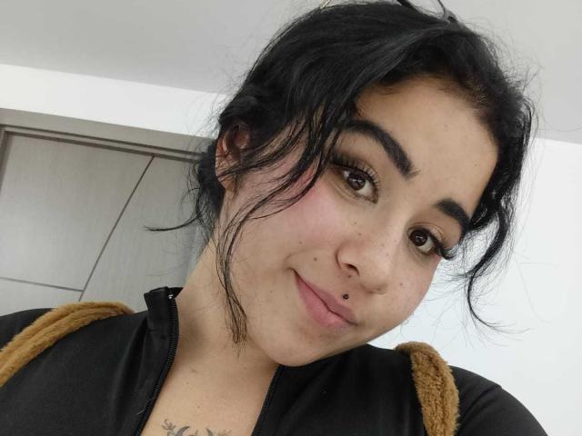 Dayanna, com cabelos pretos desalinhados, olhos castanhos e um piercing no nariz, vestindo uma jaqueta preta com zíper e tiras marrons, sorrindo levemente. Sua tatuagem é visível em seu peito..