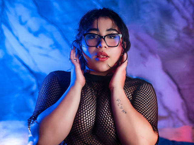 DayannaCold with black fishnet top, glasses, and hands on neck, has full tits, dark hair, tattoo on right wrist, and blue-red lighting. Her face is slightly pouty.