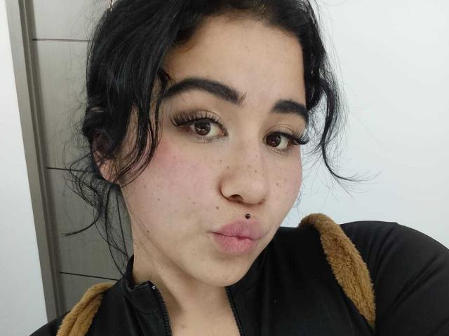 DayannaCold has black hair, brown eyes, and a small black nose piercing. She's pouting with pink lips, wearing a black shirt with tan sleeves.