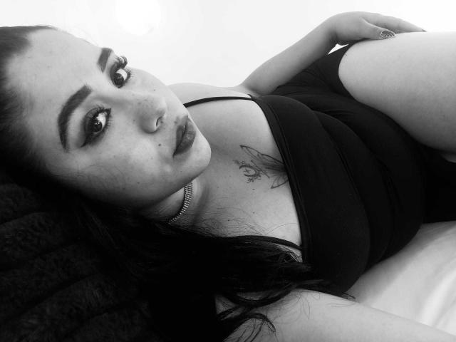 Dayanna liegt auf einem Bett, trägt ein schwarzes Tanktop, hat eine Nasenring, ein Tattoo auf der Brust, dunkles Haar, Make-up, schaut in die Kamera, eine silberne Halskette, weißer Hintergrund, Schwarz-Weiß-Foto..