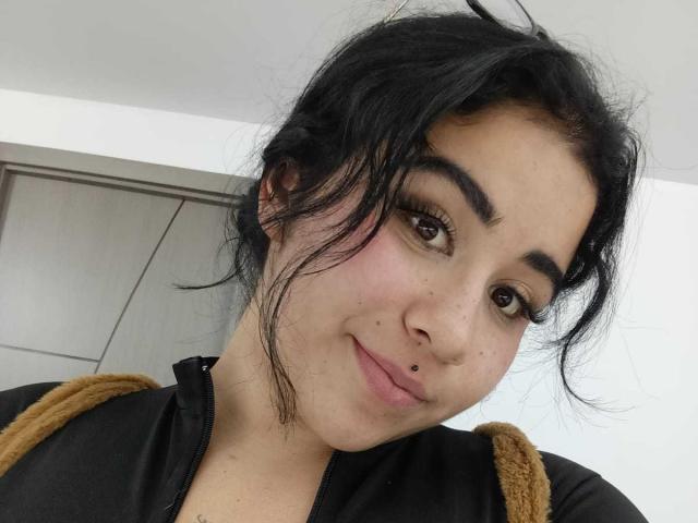 DayannaCold heeft zwart haar, bruine ogen en een kleine piercing in haar lip. Ze draagt een zwart shirt met bruine bandjes en glimlacht lichtjes. Haar neus is versierd met een klein, klein ding..