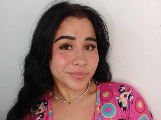 DayannaCold tem cabelos pretos longos, olhos castanhos e um sorriso discreto. Ela está vestindo uma camisa rosa com estampas de desenhos animados coloridos e um colar de pérolas. Suas nádegas não estão visíveis..