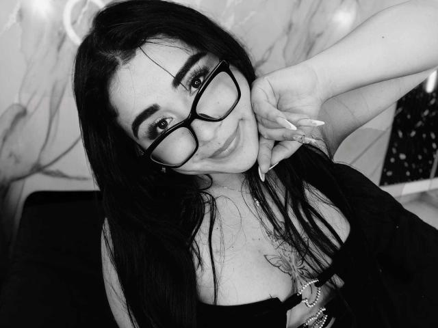 Fotografía en blanco y negro de DayannaCold con cabello largo y oscuro, gafas y una sonrisa, mostrando sus pechos y un tatuaje entre ellos, con la mano cerca de su rostro, luciendo adorable..