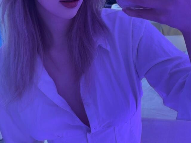 Synrix, avec un visage légèrement violet, porte une chemise blanche déboutonnée, laissant entrevoir un peu de sa poitrine, et tient un verre. Ses lèvres sont légèrement entrouvertes. Ses cheveux blonds sont visibles..