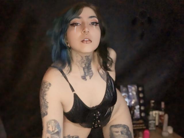 AliceCalloway with blue-black hair, black leather bra, and tattoos, has a nose ring and lip piercing, showing off her tits and tattoos, with a dark, blurry background.