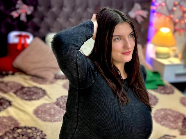 StephieRouse with dark brown hair, blue eyes, and large tits, wearing a black sweater, standing with hands in hair, smiling, in a cozy bedroom with a bed behind.