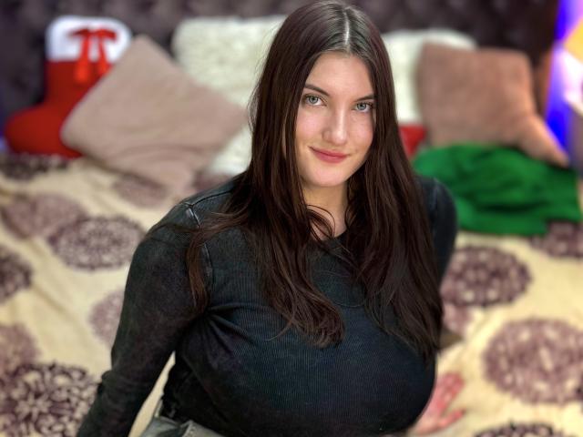 StephieRouse with long brown hair, black shirt, and big tits, smiling slightly, sitting on a bed with brown and beige patterned blanket, green and red pillows in background.