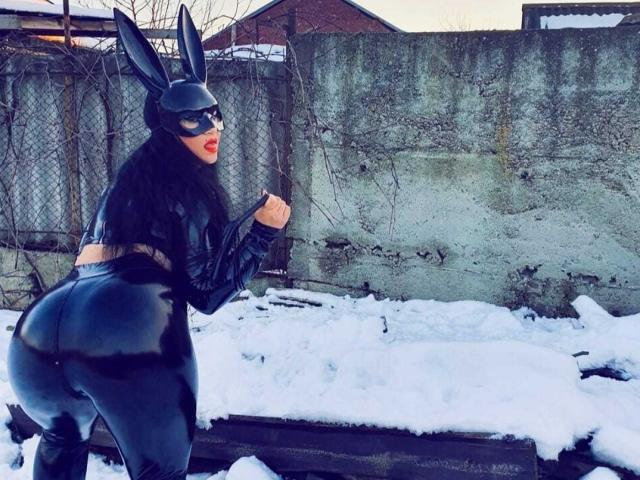 MirayDesire stands in a snowy outdoor setting, facing left. She wears a black latex bunny suit with long ears, tight around the waist and buttocks, emphasizing her curvy, large buttocks. Her long black hair is visible, and she has red lipstick. She holds a section of her suit with her right hand. The background includes a chain-link fence and a concrete wall. The image has a slightly grainy texture. Slang: "bimbo" suit.