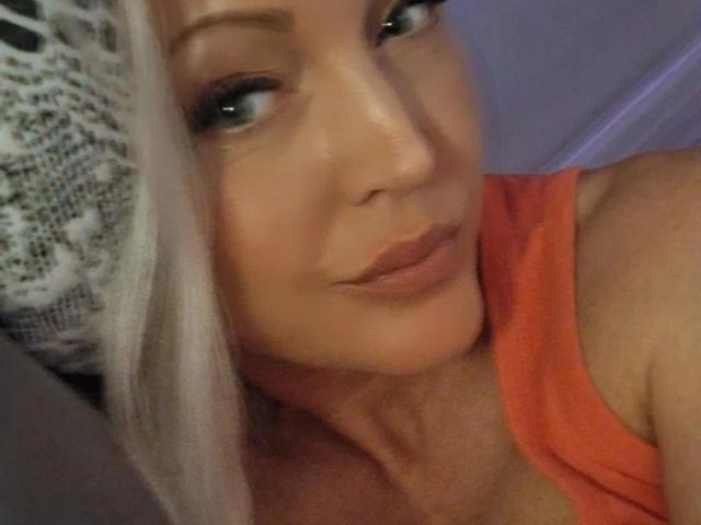 KimLayne-hot with platinum blonde hair, wearing an orange top, has full lips and green eyes, looking slightly to the side with a subtle smile. Boobs visible in the top.