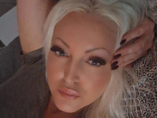 KimLayne-hot has platinum blonde hair, dark eyeliner, glossy pink lips, and is lying down with one hand on her head. She has dark nails and a gray shirt.