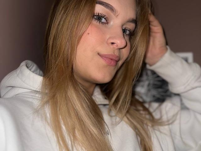 AlinaLemon has blonde hair, blue eyes, and a subtle smile. She's wearing a white hoodie, with her hand in her hair. Her nose is slightly upturned.