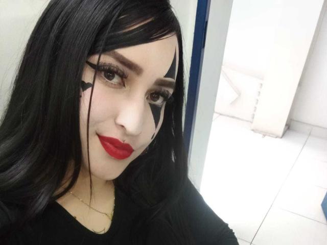 Chloey69 met lang zwart haar, rode lippenstift, een zwart gezichtsmasker en dikke wimpers, draagt een zwarte top en glimlacht naar de camera, in een kamer met witte tegels..