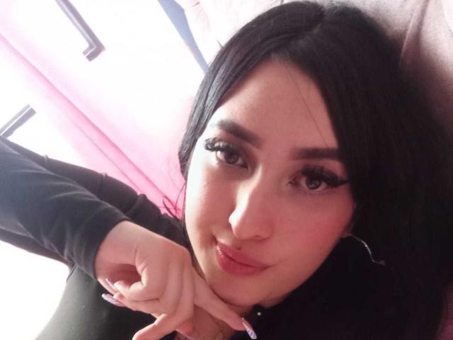 Chloey69 met donker haar, lange wimpers en een schattige glimlach, die met een vinger naar haar kin wijst, gekleed in een zwarte top, met een lichtroze huid en oorringen..