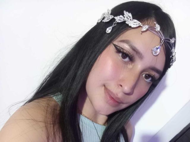 A Chloey69, com cabelos pretos longos, usando uma tiara floral prateada e uma blusa azul clara, tem delineador de olhos, um sorriso fofo e um nariz pequeno. Seus seios não estão visíveis..