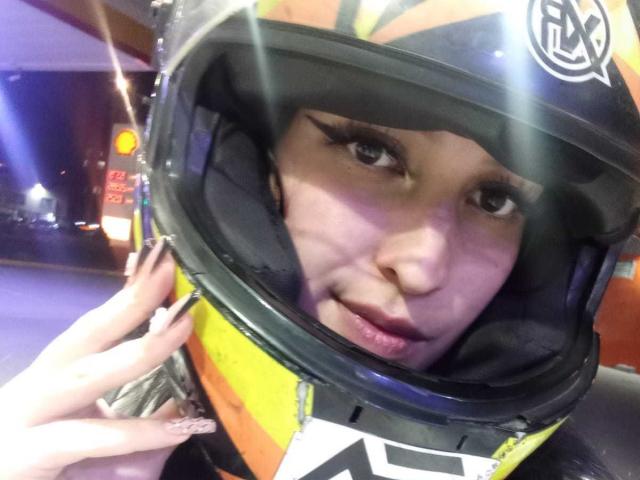 Chloey69 porte un casque de moto jaune et noir avec une visière réfléchissante, elle a de longs cils et affiche une expression légèrement boudeuse, avec sa main près du casque..