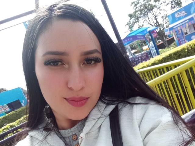 Chloey69, com cabelos pretos longos, sobrancelhas grossas e batom rosa, vestindo um moletom branco, exibindo seu grande traseiro, com um corrimão de metal amarelo ao fundo..
