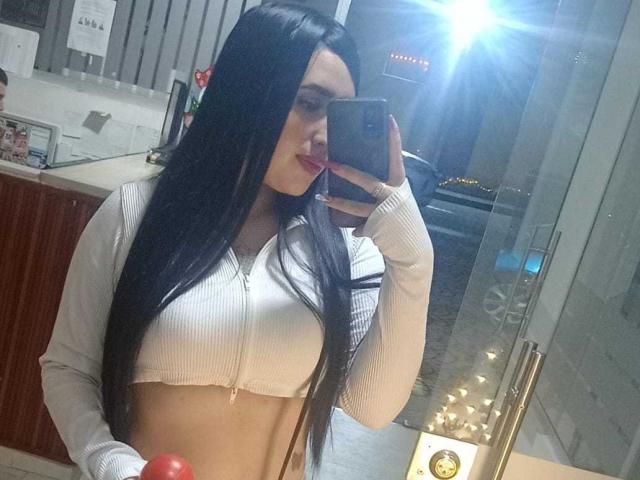 Chloey69 mit langen, schwarzen Haaren, einem weißen, gerippten Crop-Top und mittelgroßer Brust, macht ein Selfie vor einem Spiegel, ihr Handy blitzt, in einem hell erleuchteten Badezimmer..