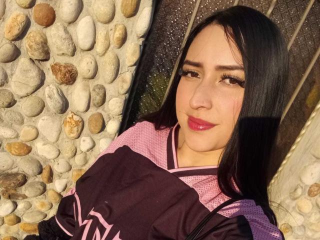 Chloey69 with long black hair, pink and purple kimono, and glossy pink lipstick, holding a dark purple bag, smiling at the camera, standing in front of a stone wall.