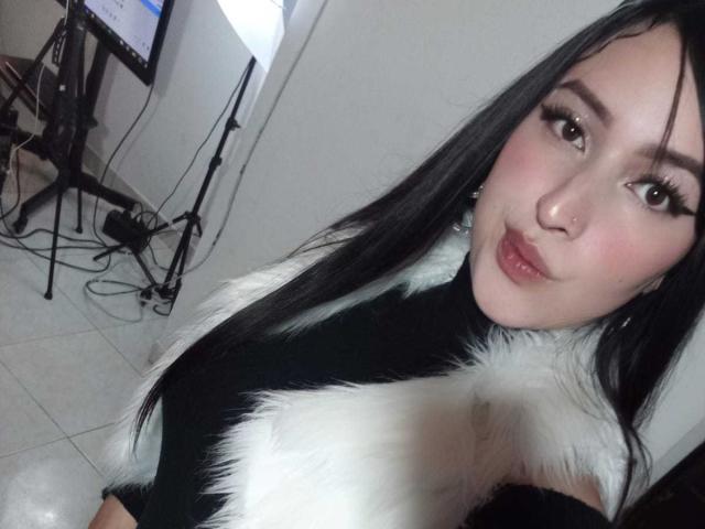 Chloey69 with long black hair, white and black fur top, pouty pink lips, nose ring, and thick eyelashes, looking at camera, with a slight smile, in a dim room.