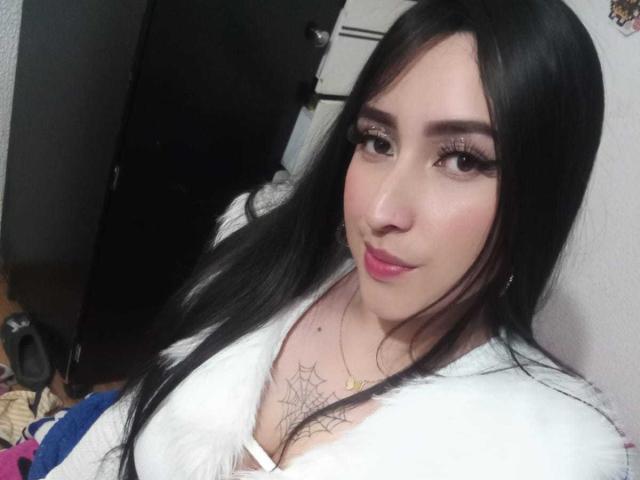 Chloey69, con lunghi capelli neri, rossetto rosa e una giacca di pelliccia bianca, mostra i suoi piccoli seni e un tatuaggio a forma di ragnatela sul petto. Ha grandi occhi e ciglia folte..