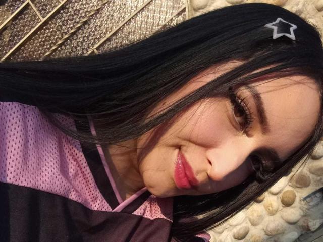 Chloey69 mit schwarzen Haaren, einem pinken und lila Hemd, einer Stern-Haarspange und pinkem Lippenstift, lächelt leicht, schaut in die Kamera und zeigt ihr süßes Gesicht und ihre glänzenden Lippen..