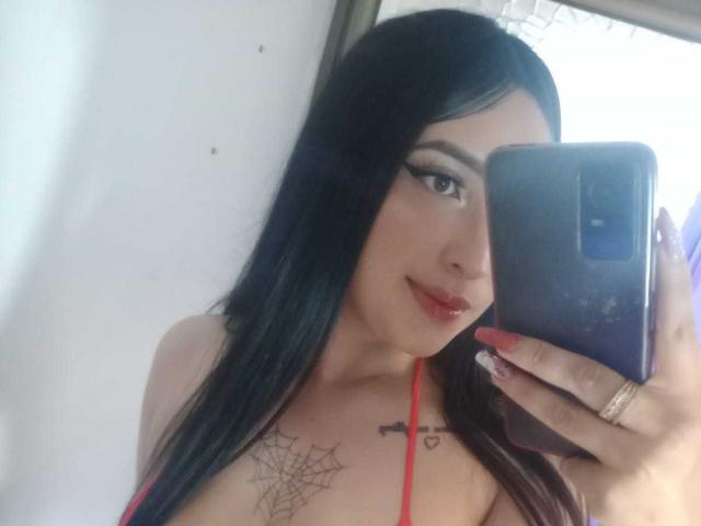 Chloey69, con i capelli neri, il rossetto rosso e un tatuaggio a forma di ragnatela sul petto, tiene in mano un telefono e mostra il seno con un top a halter rosso. Sta sorridendo leggermente..