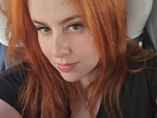 StrawberryBlond has red hair, brown eyes, and a slight smile with pink lips. She's wearing a black shirt, and her head rests on a white car seat.
