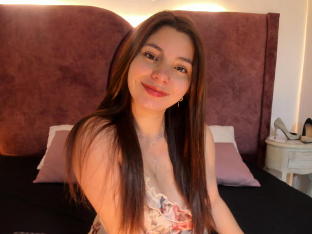LinaFoxyHot with long brown hair, smiling, wearing a floral dress, sitting on a bed, light skin, small gold earrings, bed with pink pillows and dark headboard in the background.