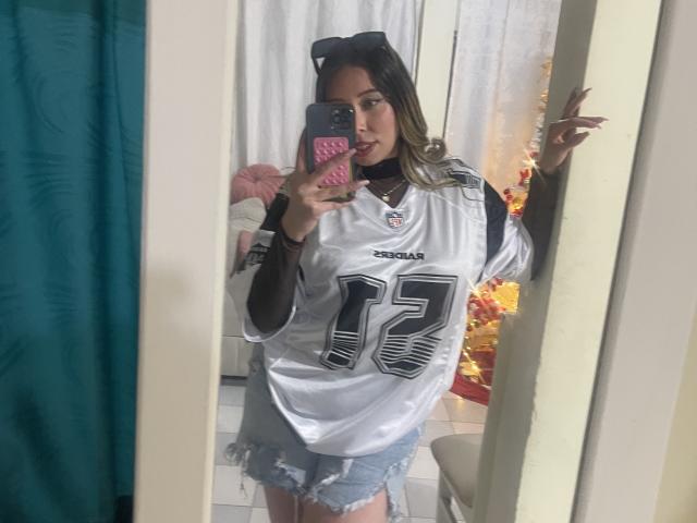 JennyLawry in a mirror selfie, wearing a white NFL jersey with number 17 and frayed denim shorts, sticking out her tongue, with a pink phone case.