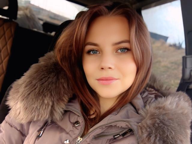 SexyVictoriaLove with blue eyes, brown hair, and pink lips, wearing a gray puffy jacket with fur-trimmed hood, sitting in a car, looking directly at the camera.