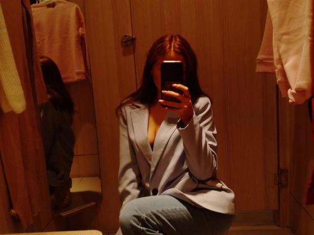 SexyVictoriaLove with red hair, sitting on floor, wearing blue jacket, and jeans, taking mirror selfie with phone, covering her face, pink towels hanging in background.