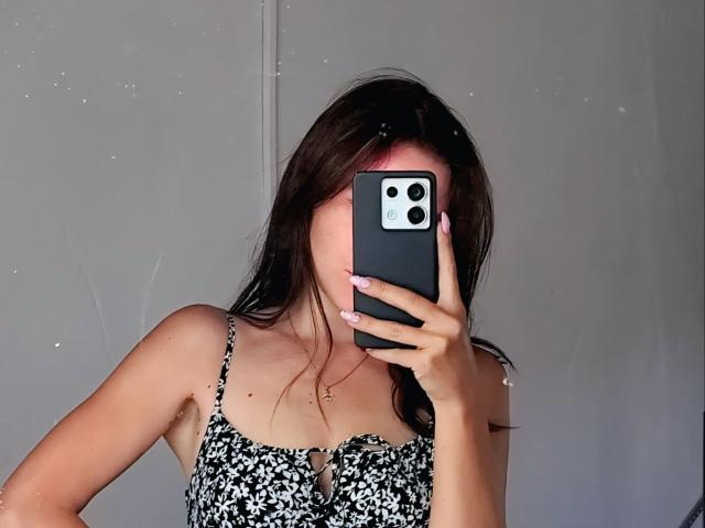 SexyVictoriaLove with long brown hair, holding a black phone covering her face, wearing a black-and-white floral dress, showing her bare shoulders and small, cute tits.