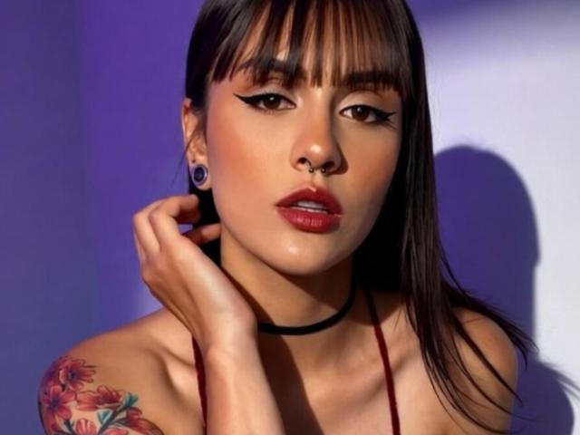MissValentine has dark brown hair with bangs, black choker, red lipstick, and a nose ring. She's touching her ear with a hand showing a colorful tattoo.