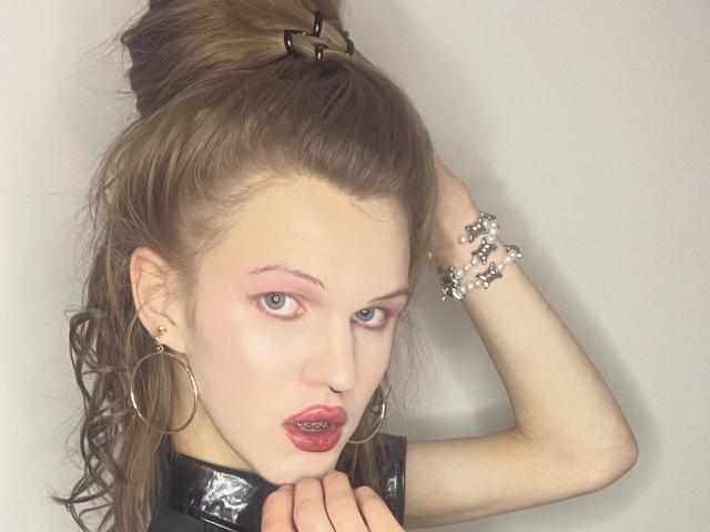 ChristyKathie with pale skin, pink eyeshadow, red lips, and hoop earrings, holding her wet brown hair up, wearing a black vinyl top, and a silver bracelet.