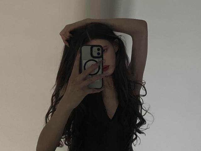 ChristyKathie with long wavy brown hair, black dress, arm raised, holding phone covering face, phone has white and black design, red lipstick, dimly lit, sexy, elegant.