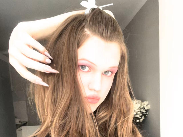 ChristyKathie with long brown hair, pink eyeshadow, and glossy pink lips, holding her hair up with one hand, showing off her nails.