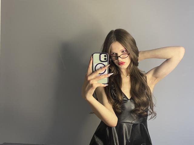 ChristyKathie with long brown hair, glasses, red lipstick, wearing a black satin dress, taking a mirror selfie, holding a phone with a white case and purple ring.