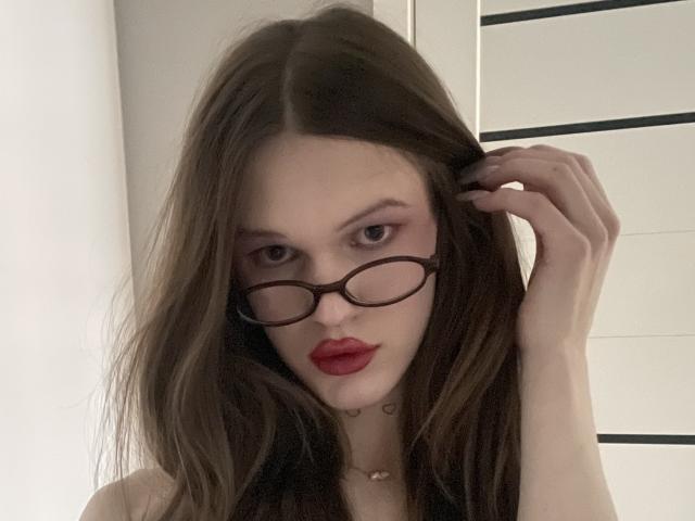 ChristyKathie with long brown hair, red lipstick, and oval glasses, holding her hair, looking at the camera, pale skin, small necklace, and a slightly annoyed expression.