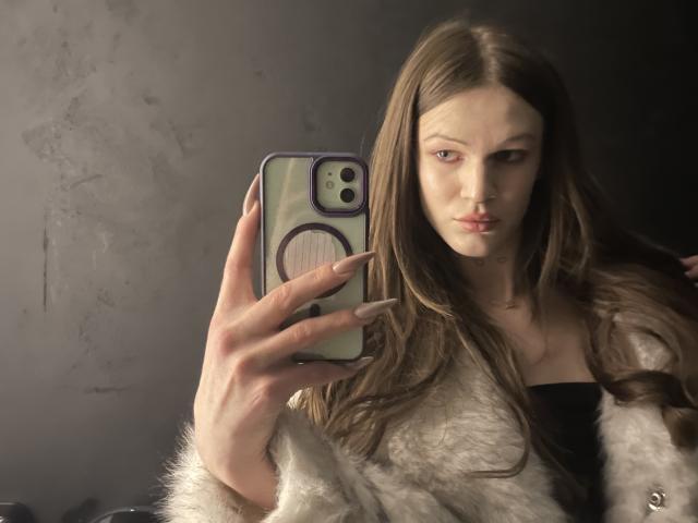 ChristyKathie with long brown hair, holding a phone with a purple ring, wearing a fluffy white jacket, and a black top, showing off her tits in the mirror selfie.