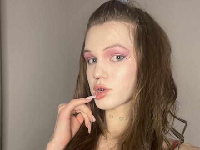 ChristyKathie with pink eye makeup, biting her finger, has wavy brown hair, pale skin, and small heart tattoos on her neck, wearing a nose ring, in a selfie.