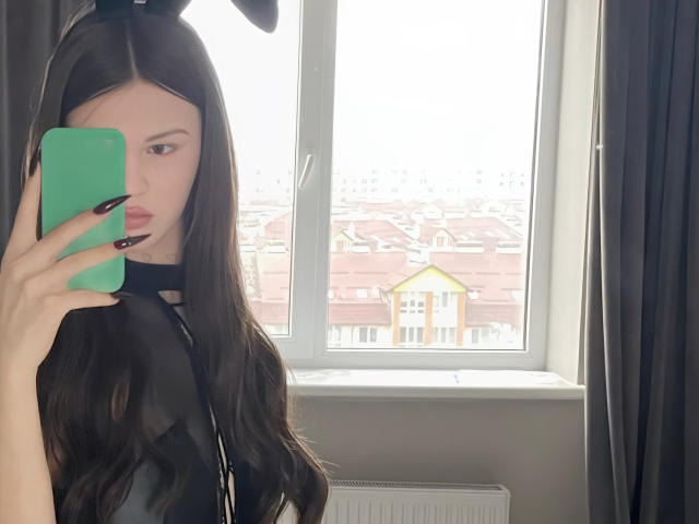ChristyKathie with long brown hair, black bunny ears, and black nails, holding a green phone, looking in a mirror, wearing a black top, showing her tits.