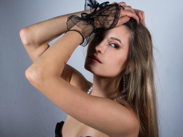 CatrinLips with long brown hair, wearing black lace glove, hands raised, showing off bare shoulders and a pearl necklace, looking at camera with slight smile.