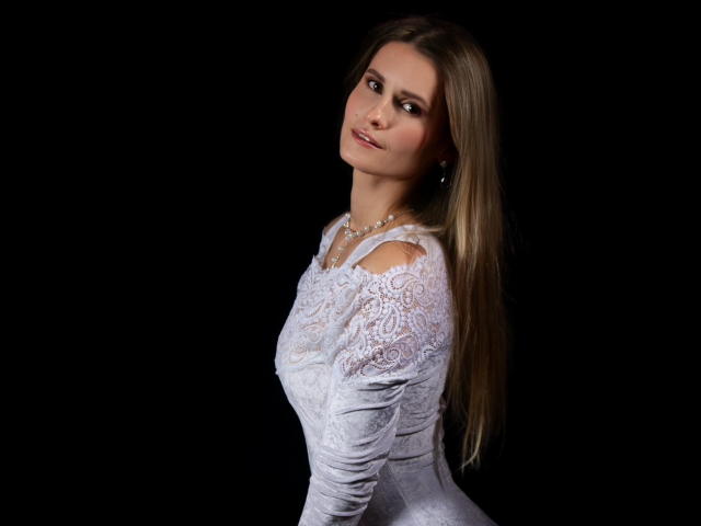 CatrinLips with long blonde hair, wearing a white lace dress, looking over shoulder with a slight smile, dark background, small pearl necklace, soft lighting.