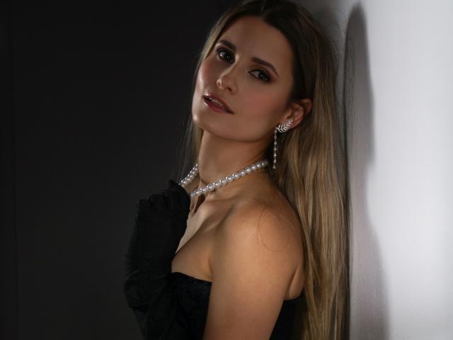 CatrinLips with long blonde hair, wearing a black strapless dress, pearl necklace, and earrings, leaning against a white wall, looking at the camera with a slight smile.
