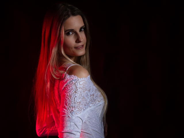 CatrinLips with long blonde hair, white lace top, red light on left, dark background, looking over shoulder, subtle smile, light skin, slim build, sexy.