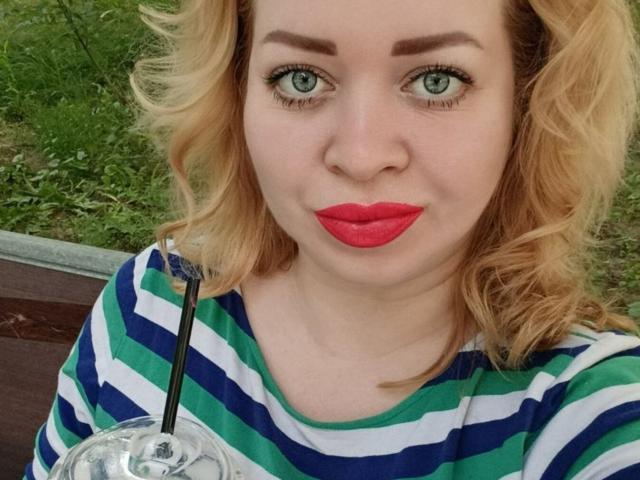 OliviaVerr, avec ses cheveux blonds bouclés, ses yeux verts, son rouge à lèvres rouge, sa chemise rayée vert-bleu-blanc, tenant une boisson claire avec une paille noire, assise à l'extérieur..