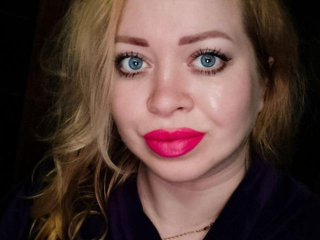 OliviaVerr has bright blue eyes, pink lipstick, and blonde hair, with a slight smile, wearing a dark purple top, against a dark background.