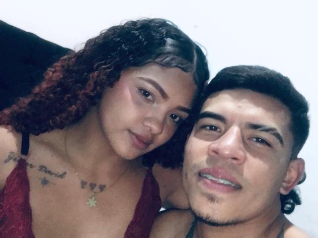 DirtyCoupleXy with curly brown hair and red dress, showing tattoos, leans on DirtyCoupleXy with short black hair and goatee, smiling at camera. Both have tan skin and earrings.