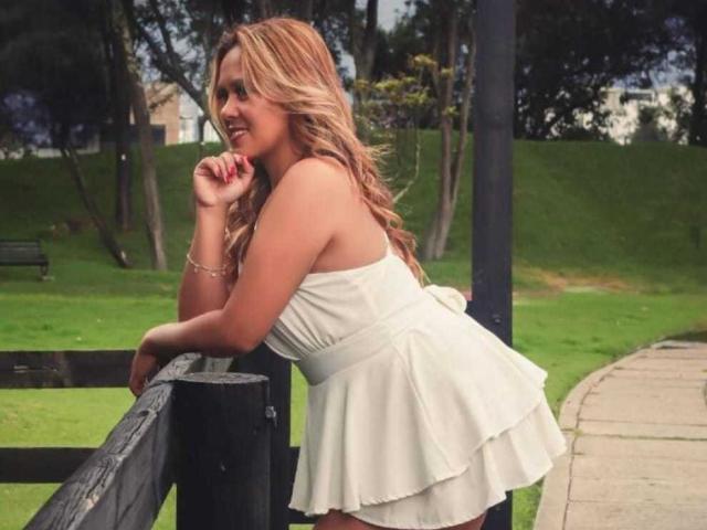 LilithBonneth with wavy blonde hair, white strapless dress, and ruffled skirt, leaning on a black fence, smiling, showing her tits, in a green park.
